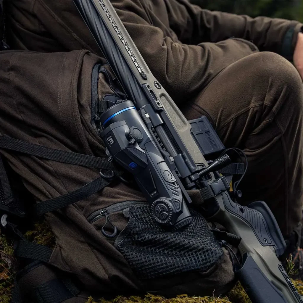 Θερμική διόπτρα Pulsar Trail 3 XQ50 τοποθετημένη σε τυφέκιο, δίπλα σε σακίδιο κυνηγού καθισμένου στο δάσος.